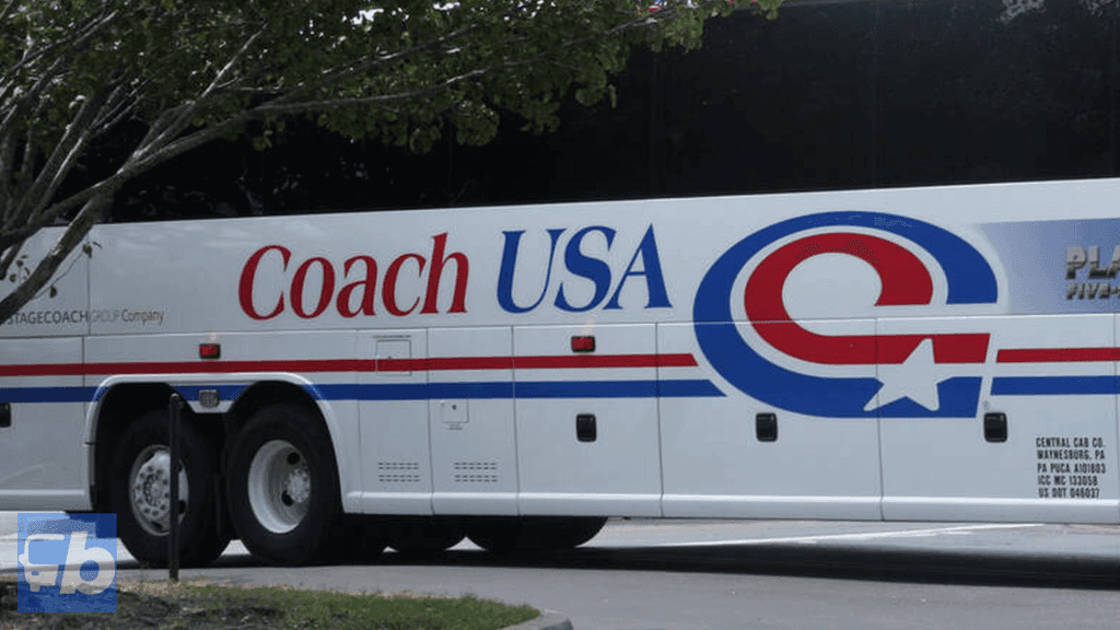 A side view of a Coach USA bus, showcasing the company's branding prominently displayed on the bus's white exterior. The logo features the name "Coach USA" in red and blue letters, accompanied by a stylized "C" with a star embedded, resembling the American flag. The bus is parked near a curb with some greenery and a tree branch partially visible in the foreground.