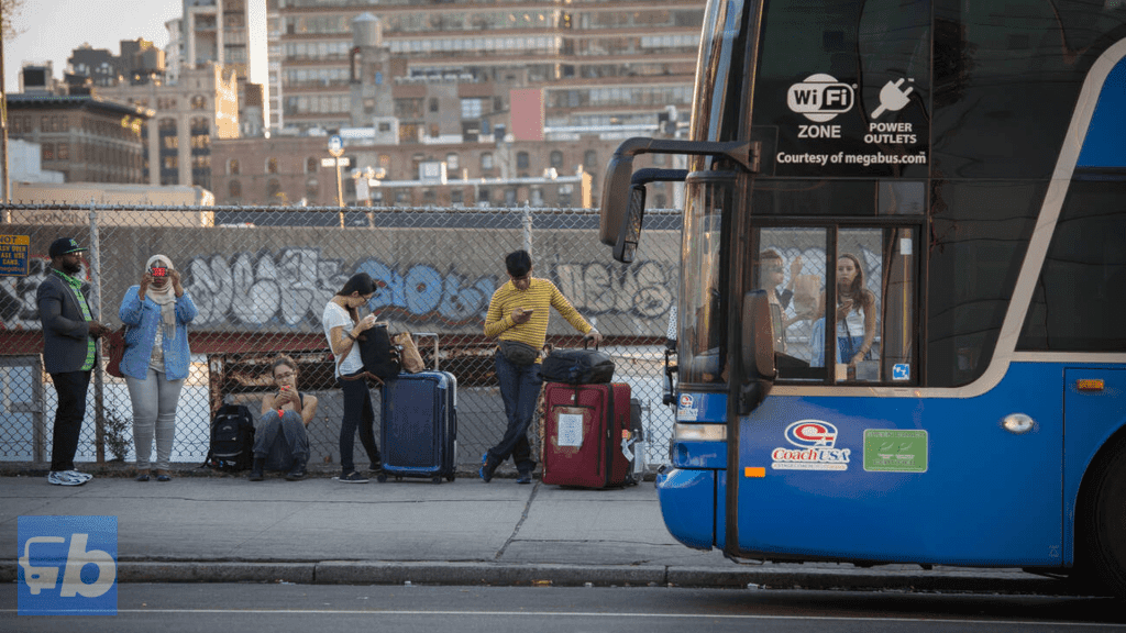 A group of passengers waiting with their luggage beside a Coach USA bus operated by Megabus at an urban bus stop. The scene includes a backdrop of city buildings and graffiti-covered fencing, creating a contrast between the organized bus service and the gritty urban environment. Some passengers are seen using their phones, indicating a modern, connected experience while waiting for the bus.