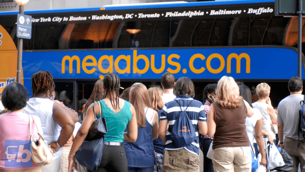 A Coach USA operated by Megabus. A large group of people is standing in line, waiting to board a Megabus at Penn Station. The bus features the recognizable blue and yellow "megabus.com" branding, with route information such as "New York City to Boston," "Washington, DC," "Toronto," and other cities displayed above the windows. The scene captures the bustling atmosphere of an urban transit hub, emphasizing the popularity and accessibility of Megabus services for travelers.
