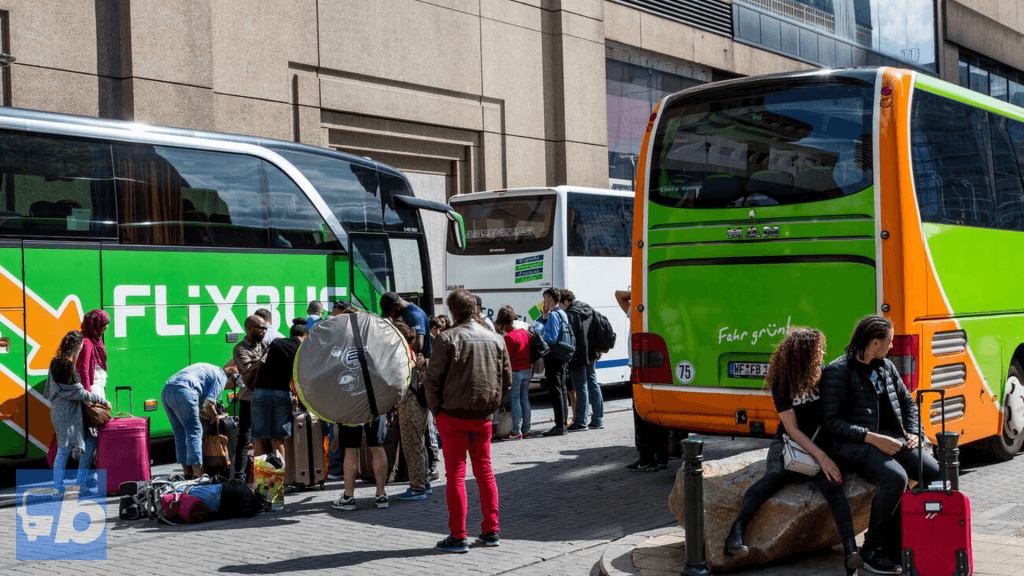A group of travelers loading their luggage into the storage compartment of a FlixBus. The bus's distinctive green and orange branding is prominently displayed, and passengers are shown in various stages of boarding.
