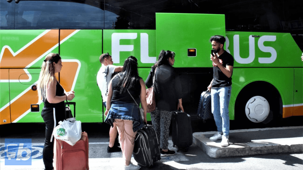A group of passengers is seen loading their luggage into the storage compartment of a FlixBus. The bus is easily recognisable with its vibrant green and orange branding, featuring a large arrow design. The passengers are holding suitcases and bags, with one individual speaking on a phone, highlighting the convenience and modern travel experience offered by FlixBus. The scene suggests a busy and organised boarding process at a bus stop.