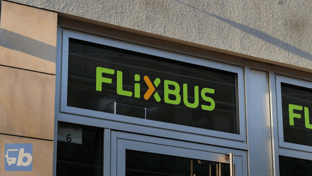 A close-up of the FlixBus ticket office sign displayed on a glass window. The bright green and orange FlixBus logo is set against a neutral-colored wall, indicating the entrance to a customer service or ticketing location.