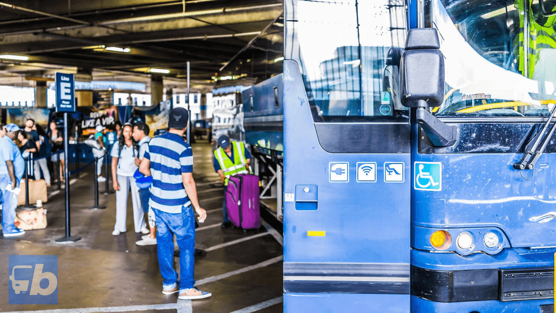 Travelers waiting in line to board a Greyhound bus at an indoor station. The bus is partially visible, and the area is equipped with signs for line organization and bus information. Wi-Fi and accessibility icons are displayed on the bus exterior.