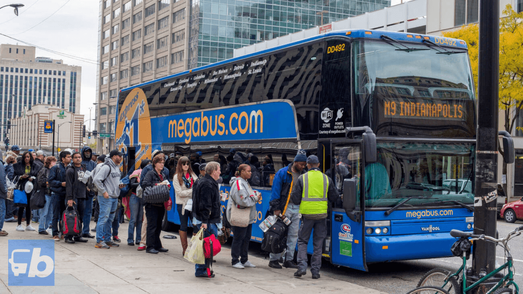Passengers lining up to board a Megabus at a city stop. The double-decker bus has 'megabus.com' written across its side in large letters. The front display indicates the bus's destination as Indianapolis. Surrounding buildings create an urban backdrop.