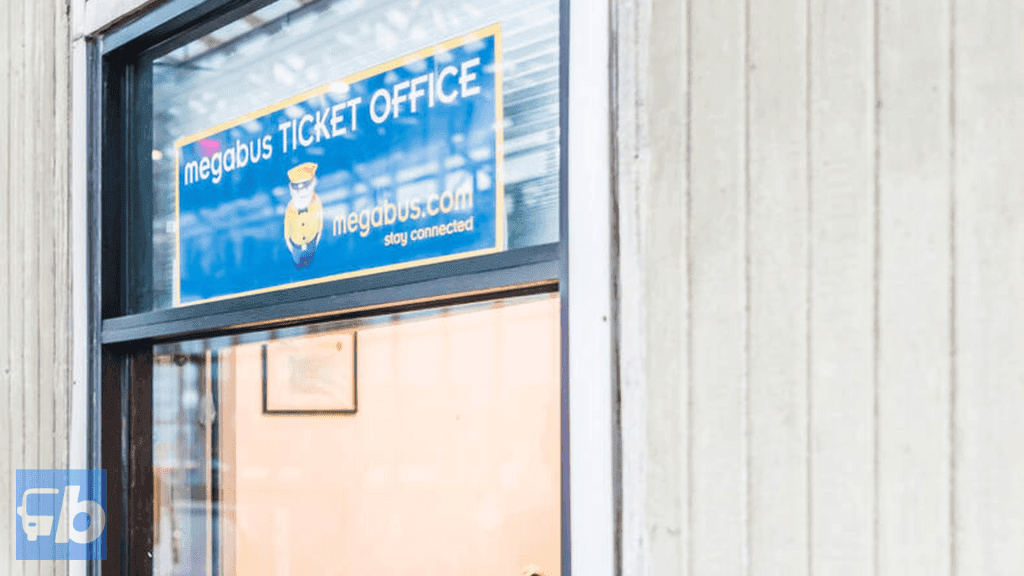 Uma vista da janela de um escritório de bilhetes Megabus, apresentando uma placa com o logotipo Megabus e o slogan "fique conectado." O escritório está em um ambiente minimalista com luz refletindo no vidro, indicando um design moderno e simplificado.