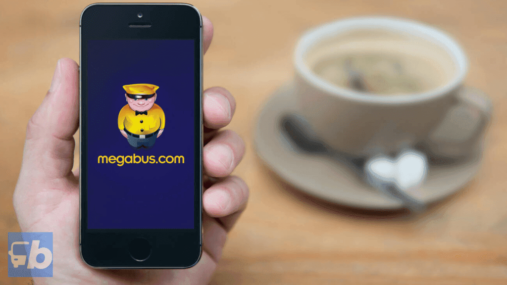 Uma mão segurando um smartphone que exibe o logotipo e a mascote do Megabus na tela. Uma xícara de café está desfocada ao fundo, sugerindo um ambiente relaxado, possivelmente enquanto se planeja ou reserva uma viagem usando o aplicativo Megabus.